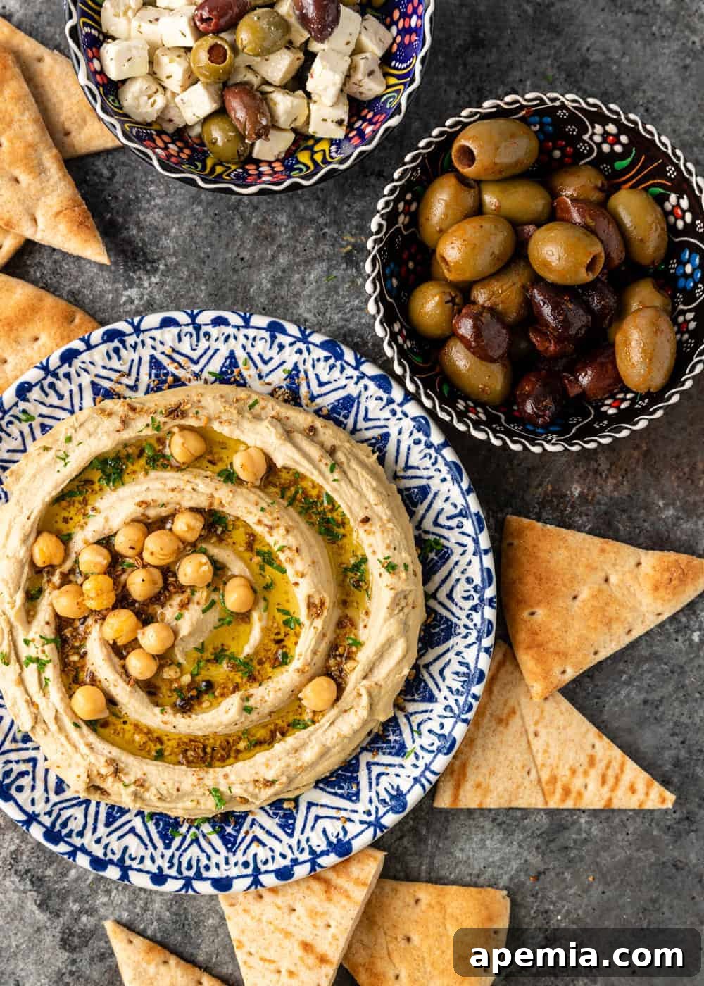 A vibrant bowl of homemade hummus, beautifully arranged with a side of golden pita chips, briny olives, and creamy cubes of feta cheese, perfect for a Mediterranean spread.