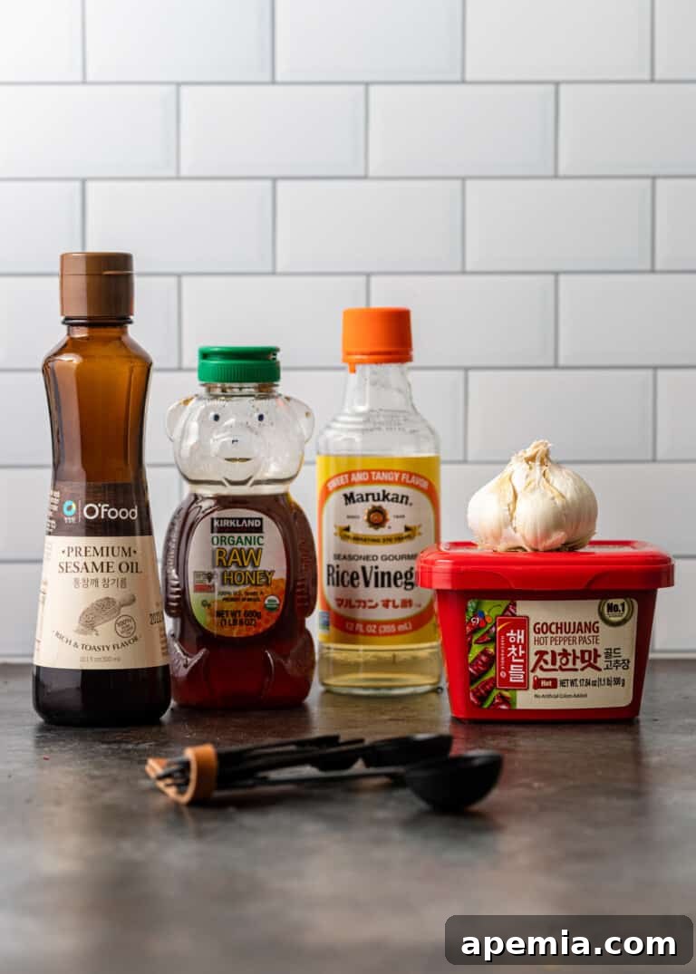 A collection of essential ingredients for Gochujang sauce including bottles of sesame oil, raw honey, rice vinegar, and a container of vibrant red chili paste.