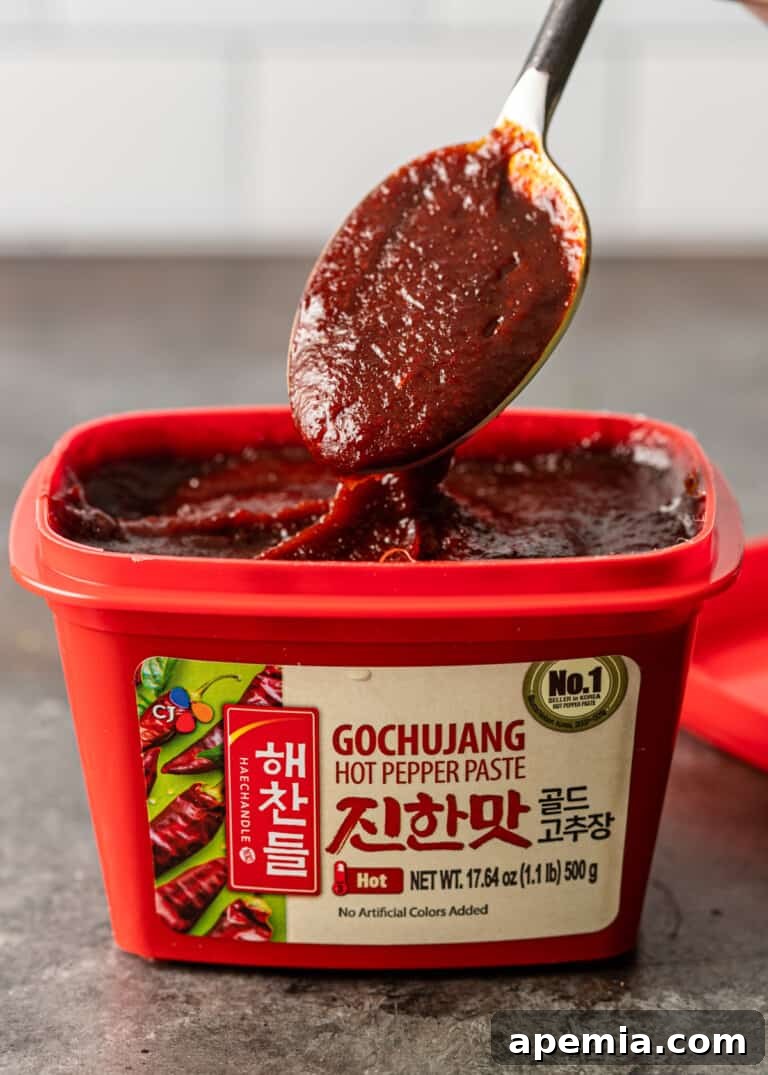 Close-up shot of a spoon scooping thick, glossy gochujang paste from its container, showcasing its rich red color and texture.