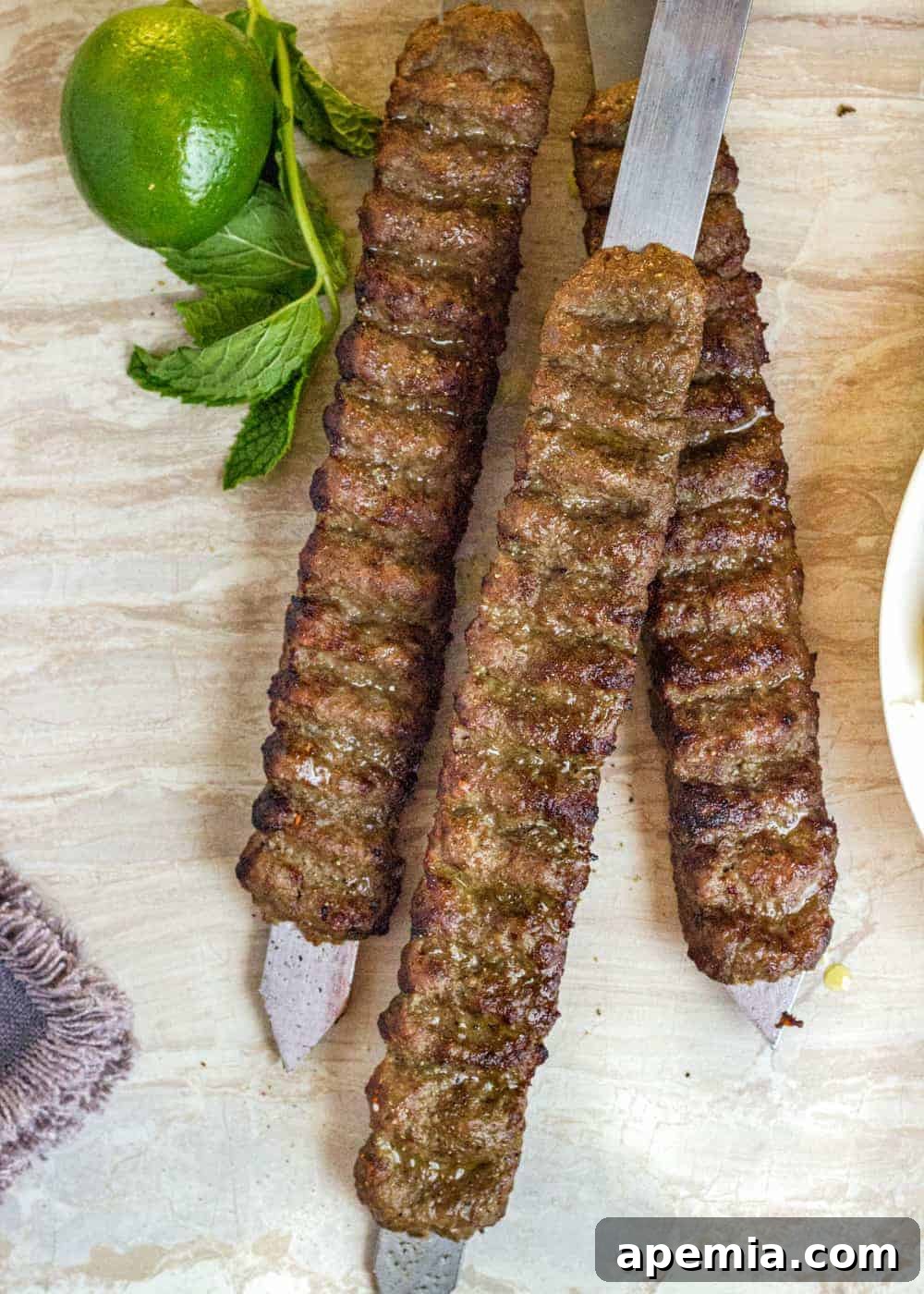 Hand-shaped Koobideh kabobs on flat metal skewers, garnished with fresh mint leaves and a whole lime, showcasing the vibrant ingredients.