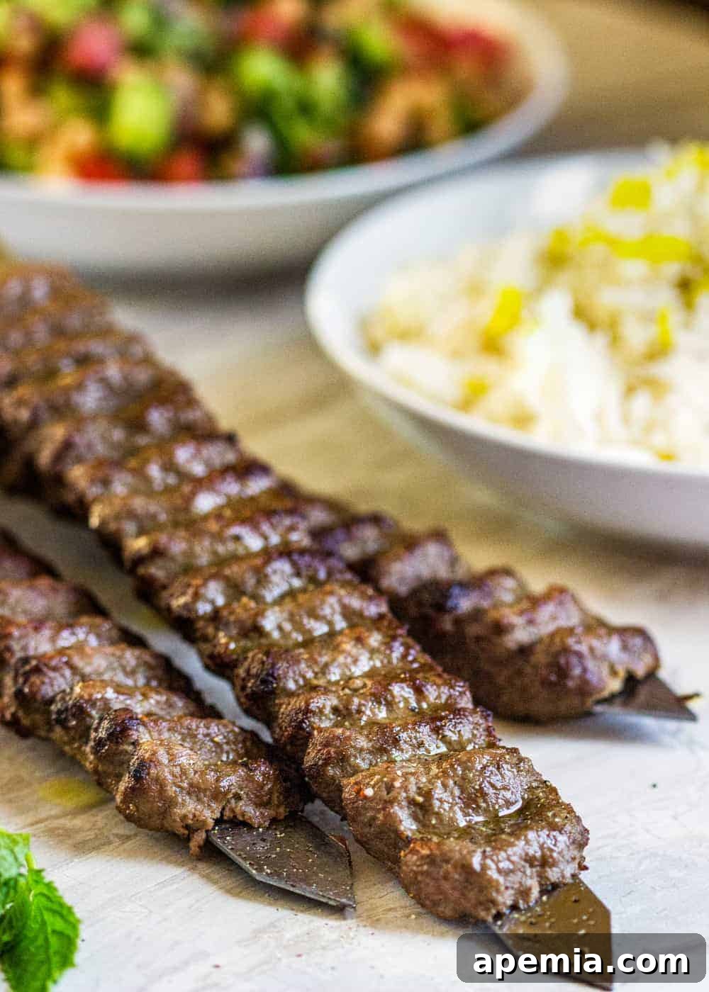 A close-up of grilled Koobideh kabobs in the foreground, with bowls of fluffy Persian saffron rice and a fresh salad blurring in the background, showcasing a complete meal.