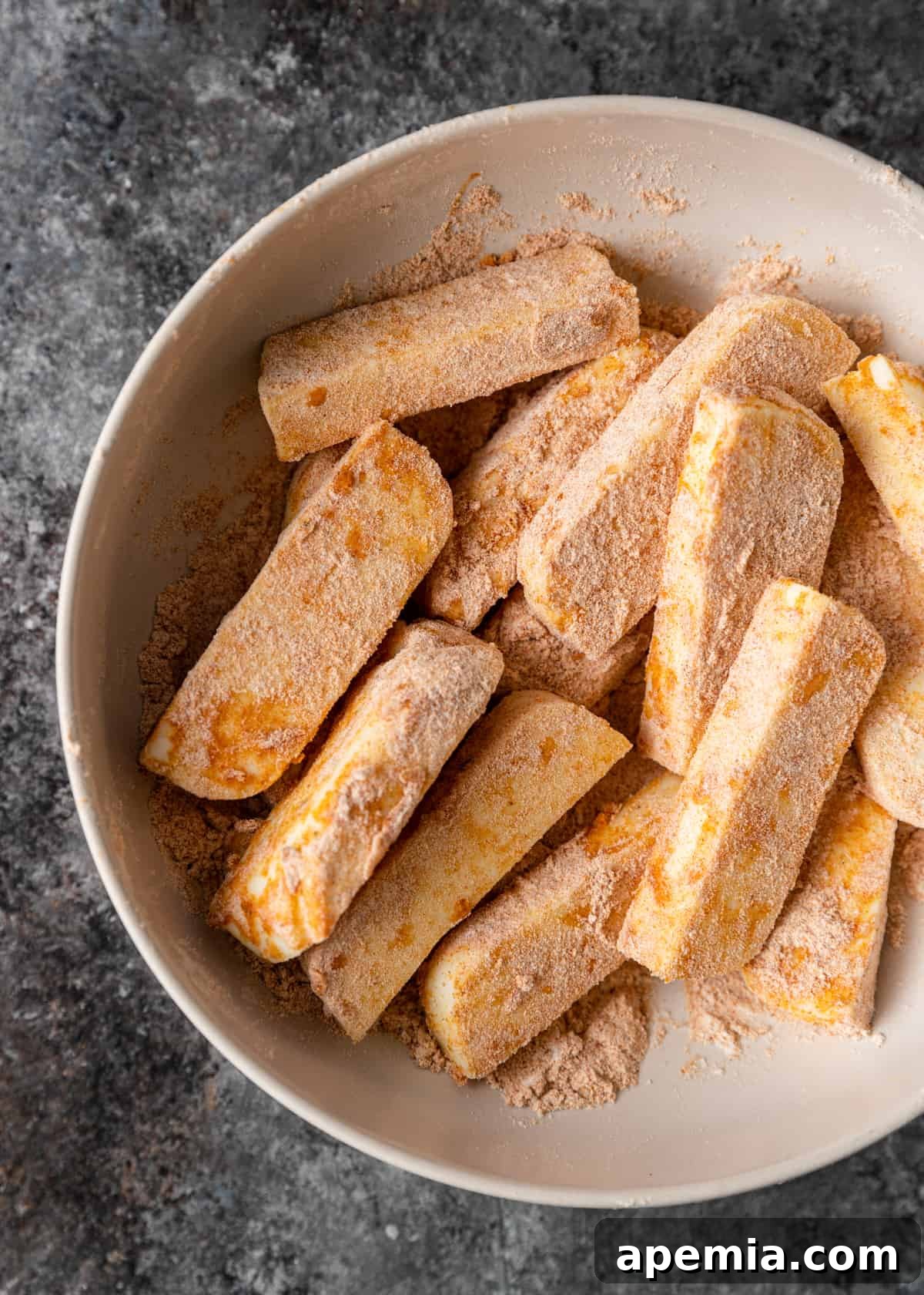 Overhead shot showing halloumi strips being coated evenly in a seasoned flour mixture in a shallow bowl.