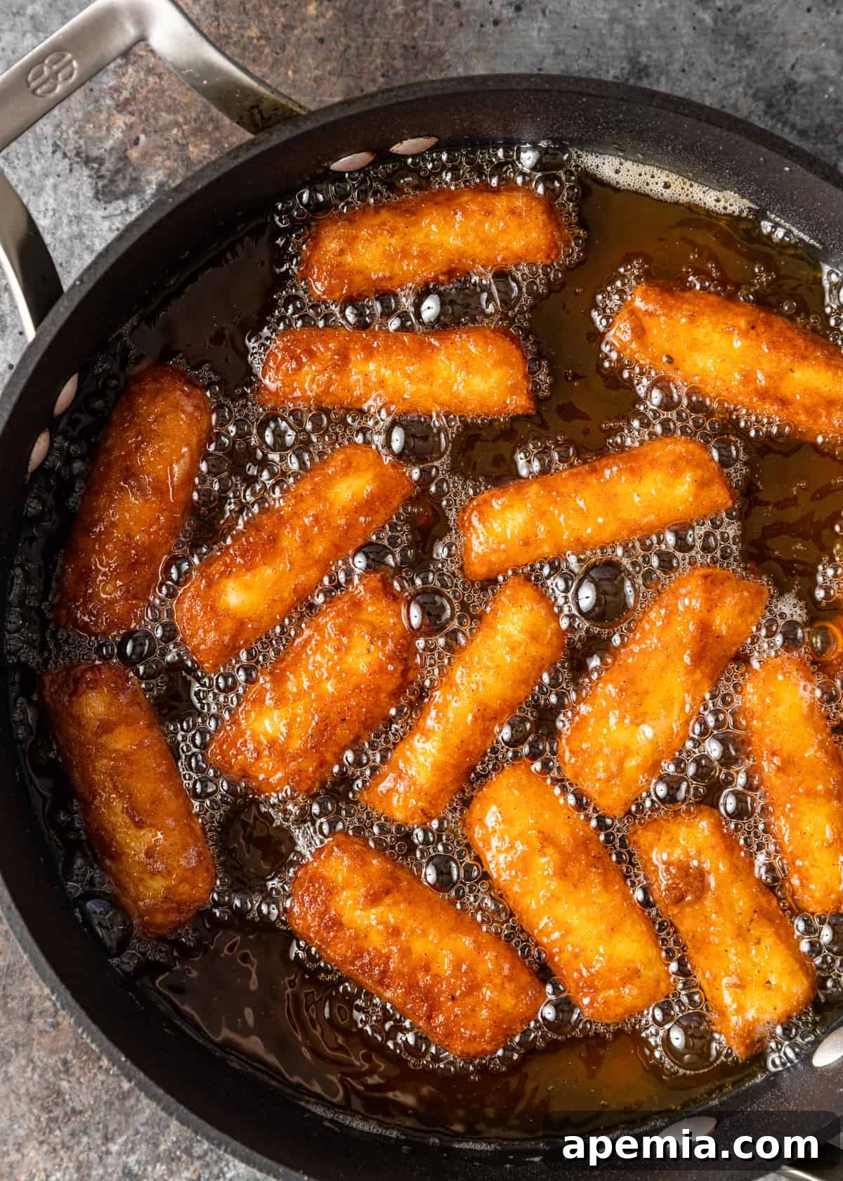 Overhead view of halloumi fries gently frying in hot oil in a deep skillet, turning golden brown.