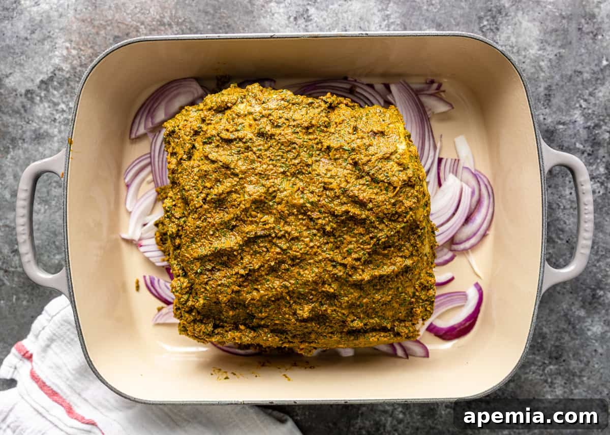Overhead process shot showing a lamb shoulder generously smothered in the rich, dark marinade, resting on a bed of sliced red onions in a roasting pan.