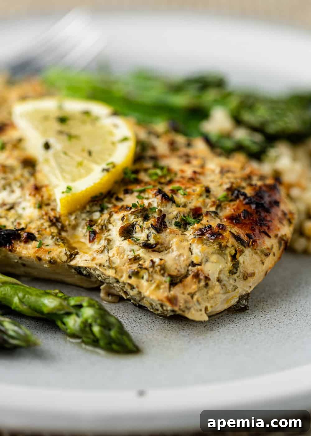 Closeup of baked Greek chicken with a vibrant slice of lemon and fresh herbs on top, served with a side of tender asparagus.