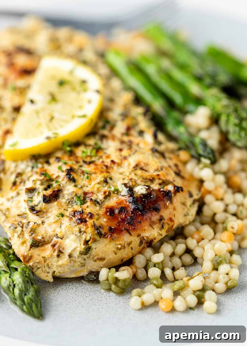 Closeup of beautifully baked Greek chicken, garnished with fresh lemon and herbs, served over a bed of fluffy couscous.