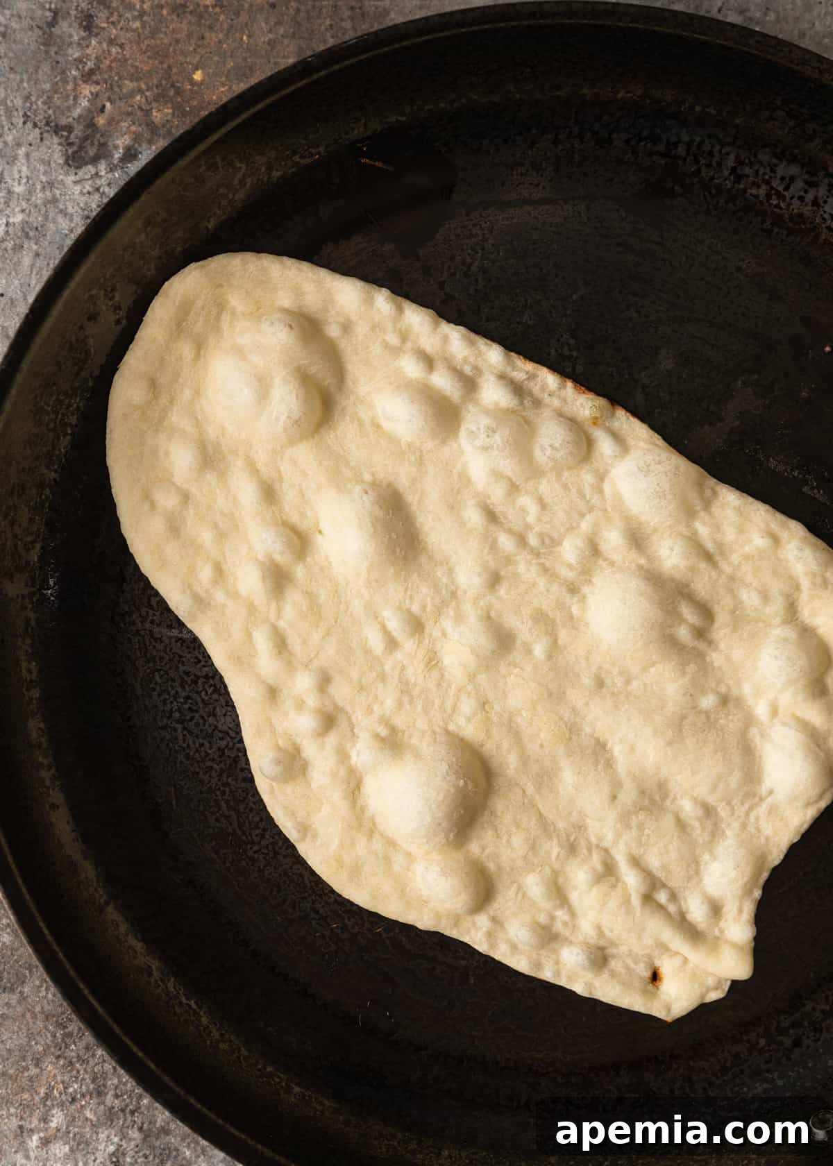 Overhead shot of naan bread cooking in a hot cast iron skillet, showing numerous bubbles forming on the surface.