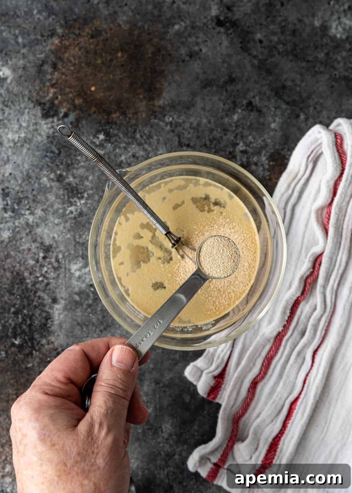 Overhead shot of dry yeast being spooned into a bowl with warm water and sugar, initiating the activation process.