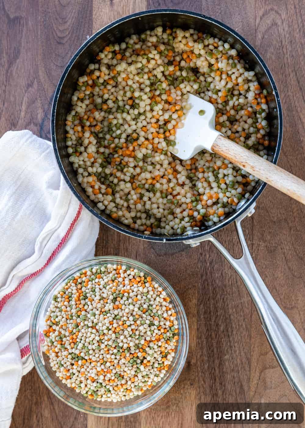 Close-up overhead view of a saucepan filled with perfectly cooked, golden pearl couscous, with a small prep bowl of uncooked couscous beside it, illustrating the before and after states.
