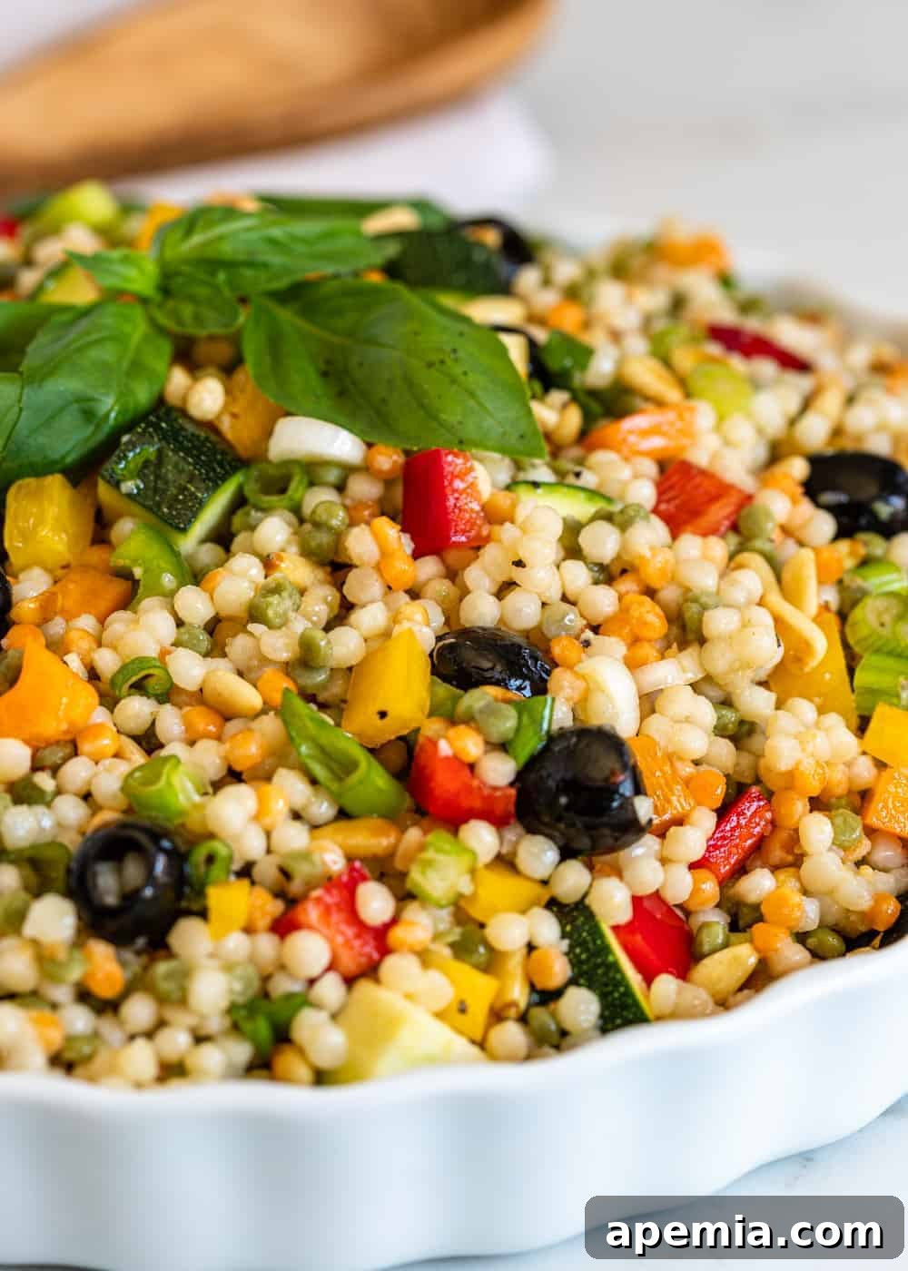 Close-up shot of a vibrant Israeli couscous salad (Ptitim) in a white serving dish, highlighting the texture of the couscous and the colorful diced vegetables and herbs.