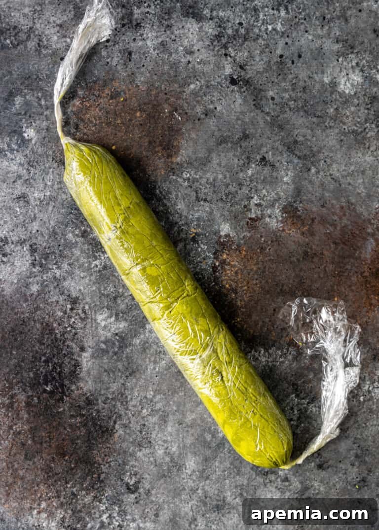 Overhead process shot: matcha cookie batter neatly rolled into a log shape and wrapped in plastic wrap, ready for chilling.