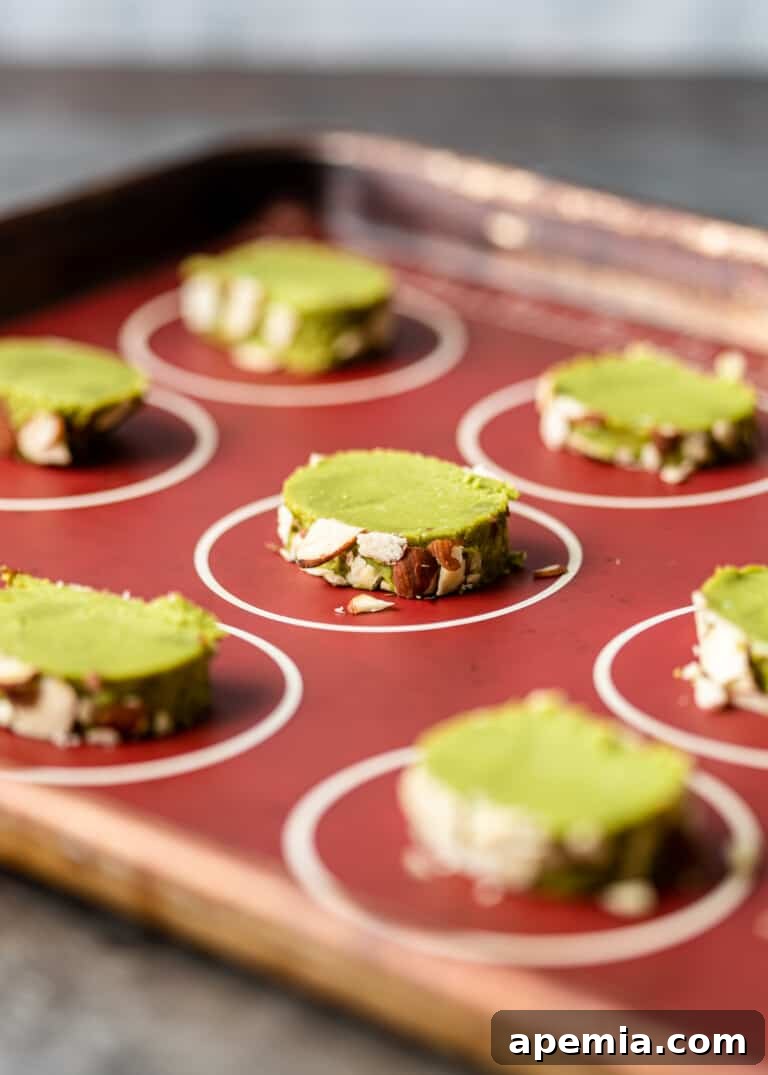 Side view process shot: unbaked matcha cookie dough slices, coated with slivered almonds, neatly arranged on a baking sheet.