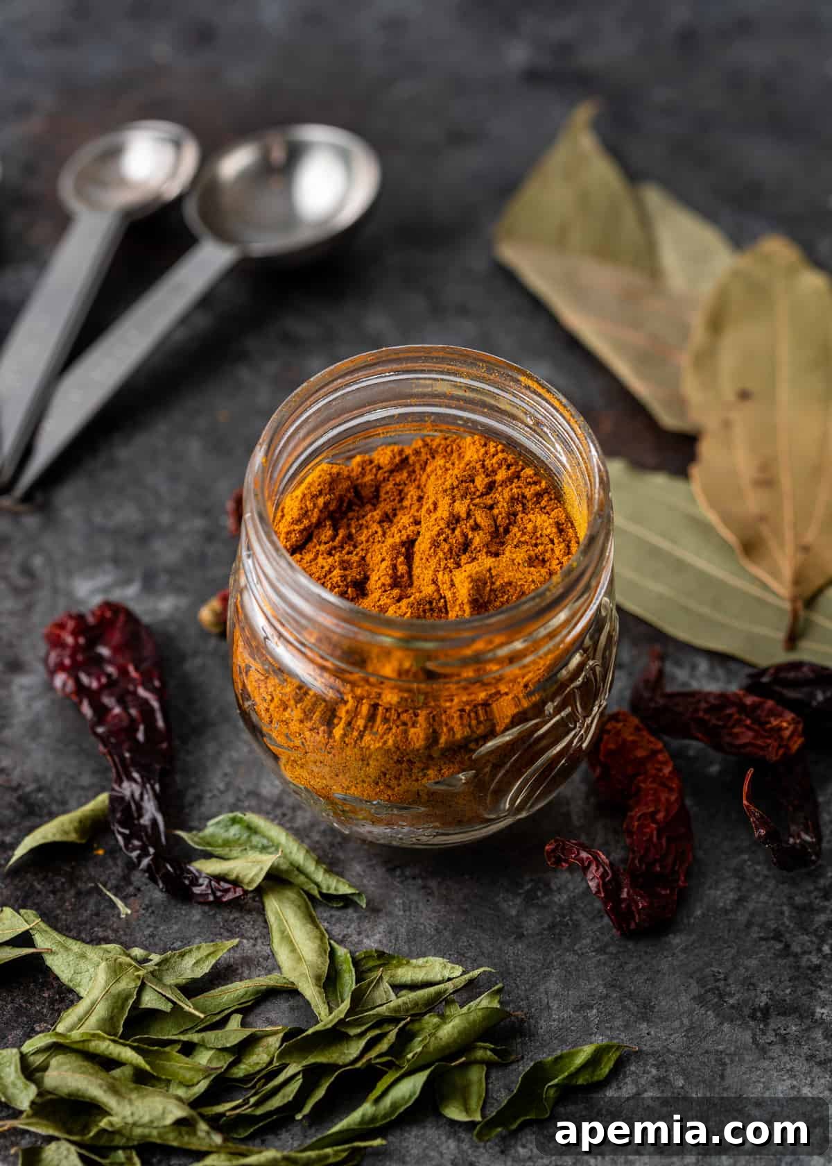 A glass jar filled with freshly made Madras Curry Powder, with dried chiles and curry leaves scattered around.