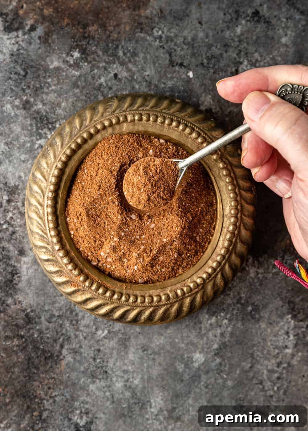 A hand holding a small, decorative golden spoon filled with the finely ground Lebanese 7 spice blend, poised over a matching golden spice dish.