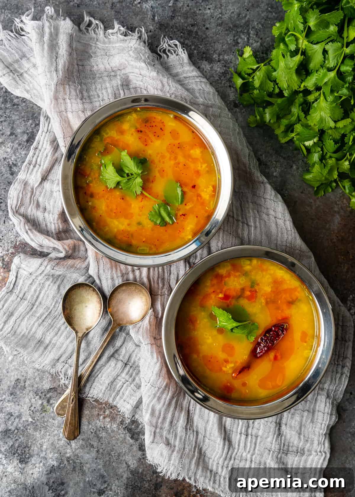 Two sleek silver bowls brimming with vibrant golden Dal Tadka, artfully garnished with fresh green cilantro, presented on a light-colored hand towel. The arrangement highlights the appealing texture and inviting presentation of the dish, ready for serving.