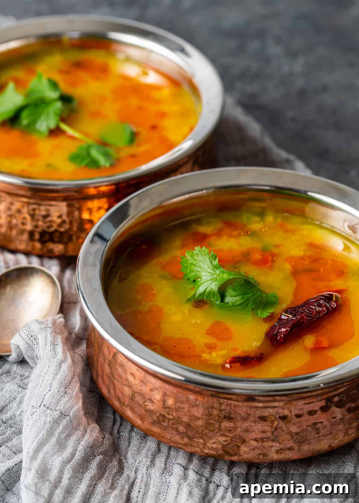 A close-up side view of a steaming bowl of Dal Tadka, showing its rich, creamy texture and the vibrant, sizzling tadka expertly drizzled on top. Garnished with fresh green cilantro, the dish looks incredibly appetizing and inviting, ready to be enjoyed.