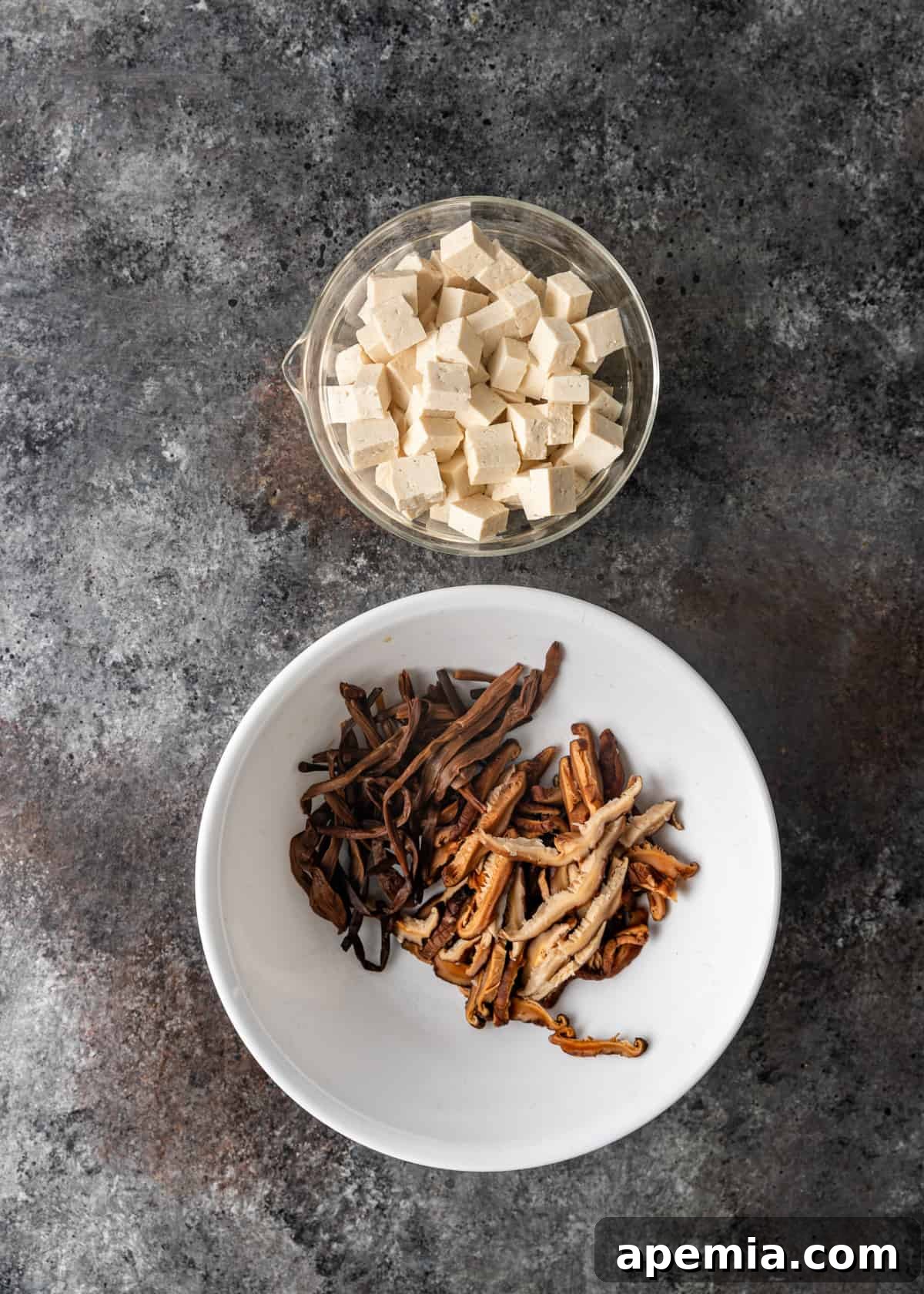 Overhead view of cubed firm tofu and rehydrated, thinly sliced dried vegetables (likely mushrooms and lily flowers) in separate bowls for hot and sour soup.