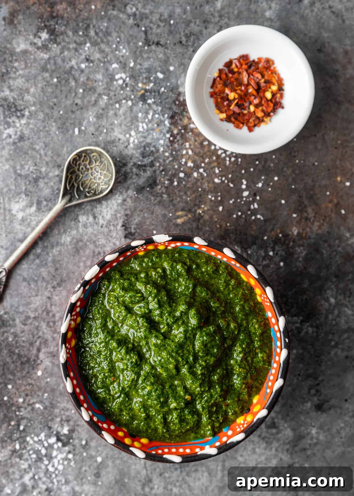 Overhead view: a small bowl of green chermoula with a bowl of red pepper flakes beside it, ready for serving.