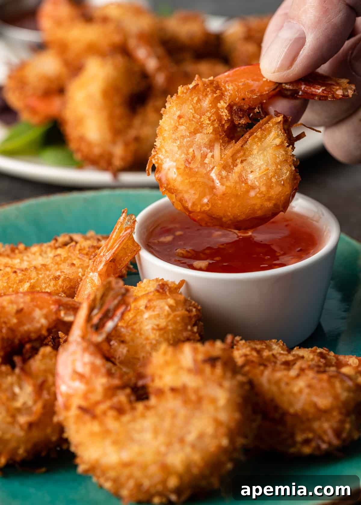 Sweet Coconut Crunch Shrimp 3 Closeup side view: a hand dips a crispy coconut shrimp into a small bowl of vibrant sweet chili sauce.
