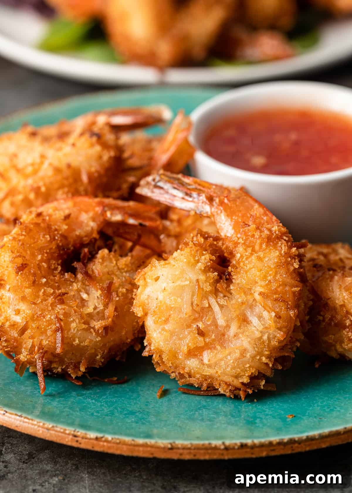 Sweet Coconut Crunch Shrimp 10 Closeup side view: a perfect pile of crispy coconut shrimp resting on a turquoise plate with a small bowl of sweet chili sauce in the background.