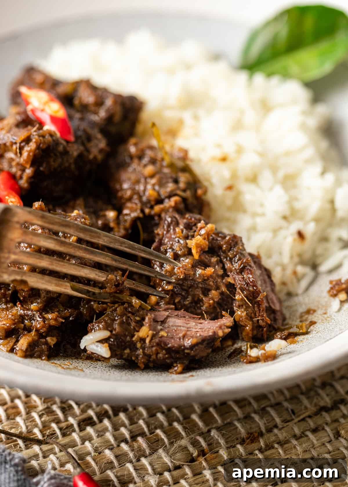 Closeup side view of a fork gently cutting into a piece of tender Beef Rendang curry on a white plate, served alongside white rice, showcasing the meat's perfect texture.