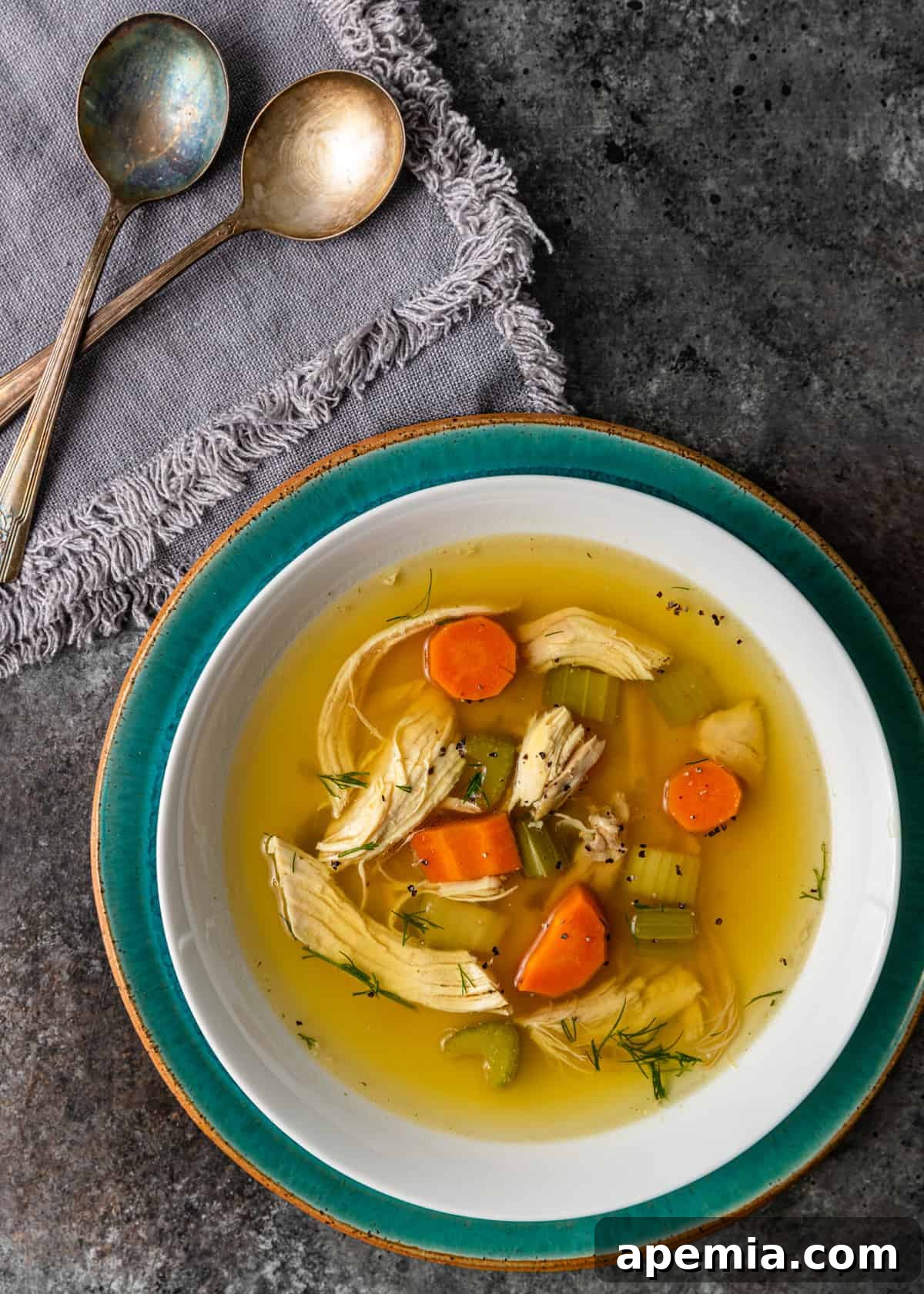overhead: matzo ball soup in a white and green bowl on top of countertop. Two spoons off to side on gray linen