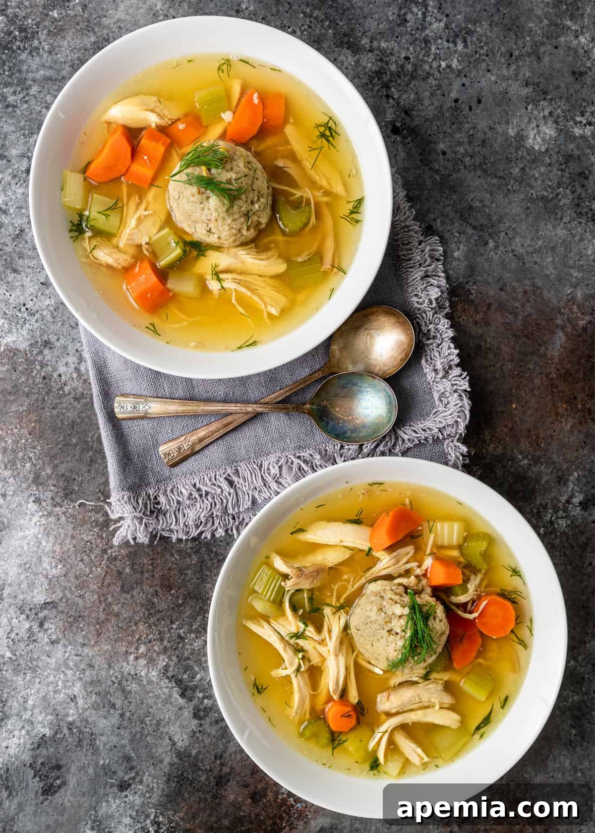 overhead: matzo ball soup in two white bowls with two spoons on gray linen