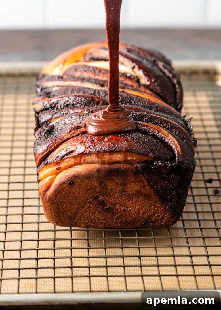 Overhead process shot: Baked babka on a wire rack with a rich chocolate glaze being poured generously over the top.
