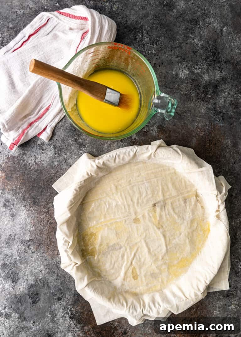 Process shot: Brushing melted butter generously onto layers of filo sheets in a pie dish, preparing the base of the pastilla.