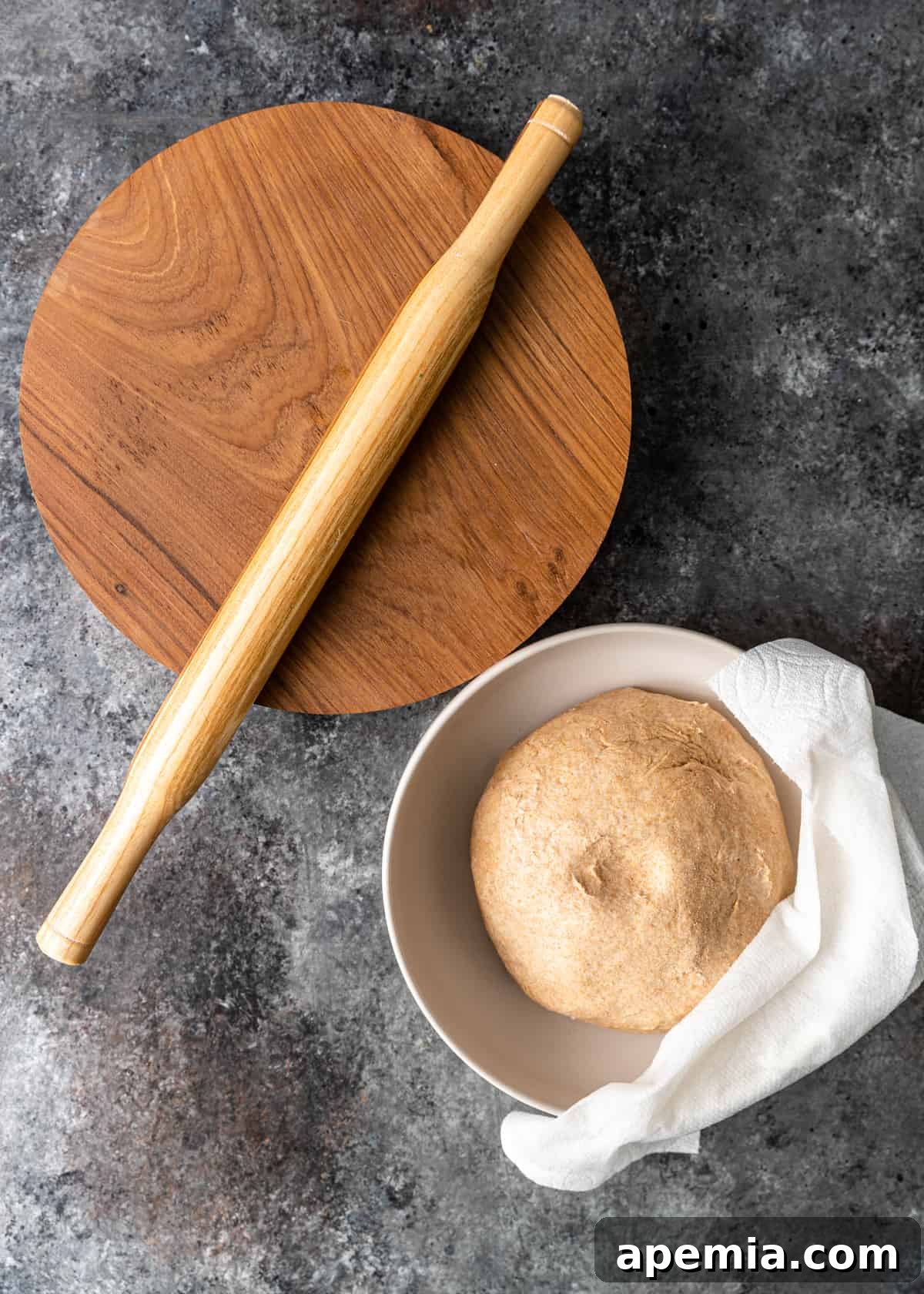 Overhead view of a rolling pin resting on a cutting board next to a white bowl filled with freshly kneaded Roti dough
