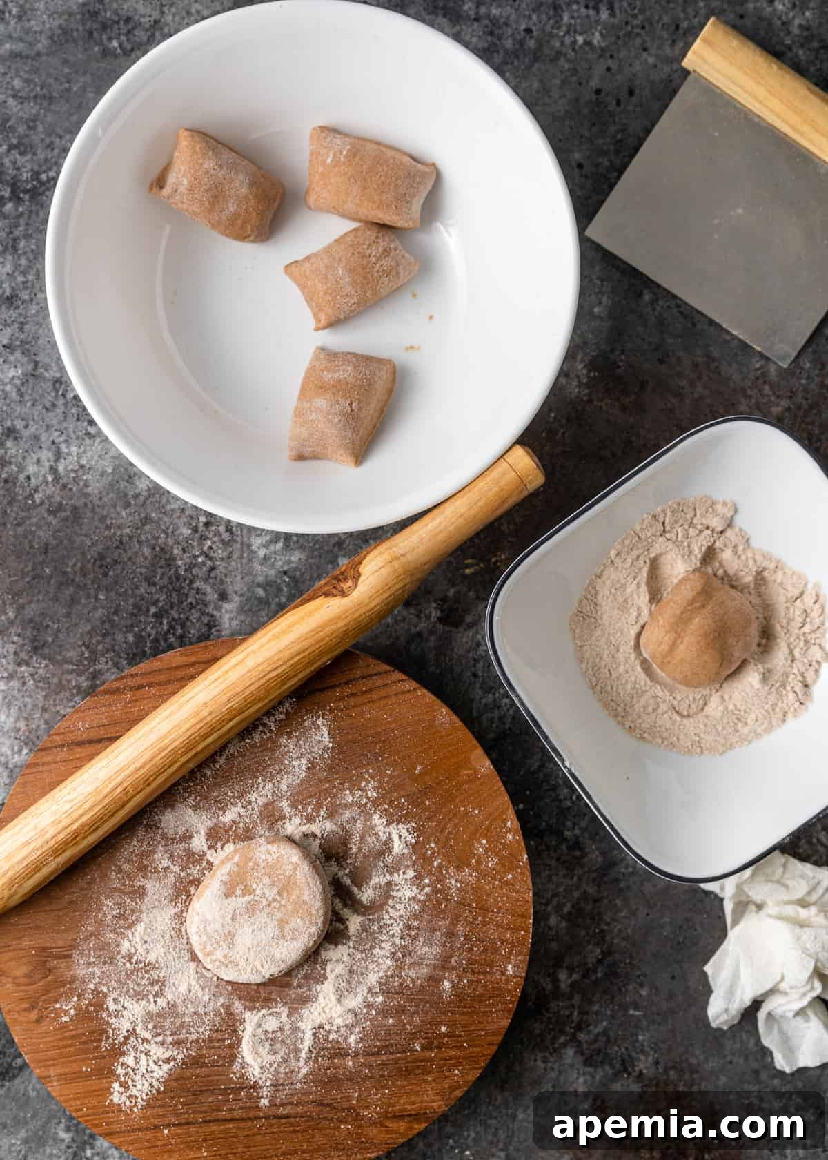 Overhead process shot illustrating two white bowls with cut pieces of Roti dough, alongside a cutting board with one piece of dough lightly sprinkled with flour and a rolling pin, preparing for rolling.