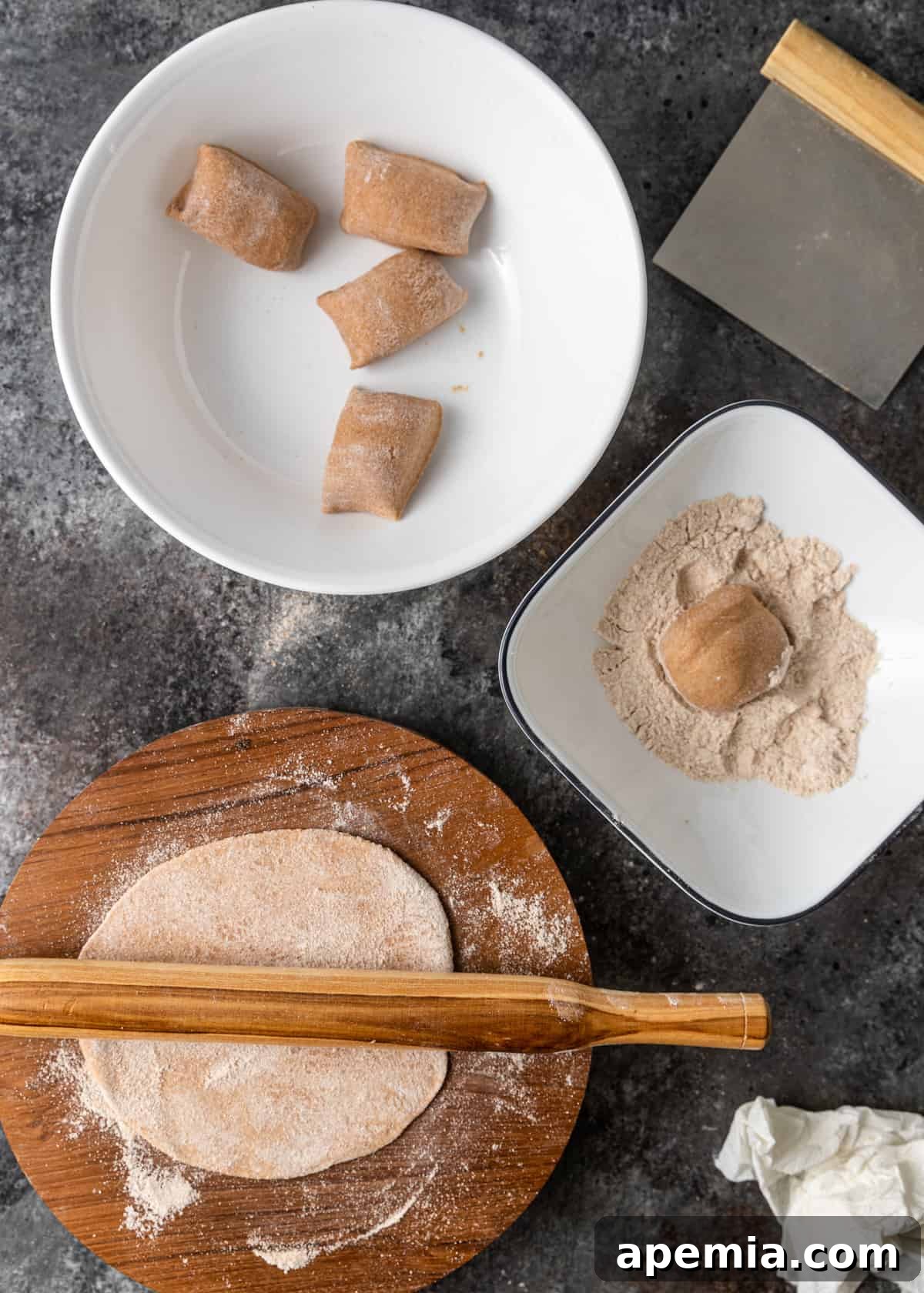 Overhead process shot capturing two white bowls of Roti dough portions, with a rolling pin actively shaping a piece of dough into a thin, round circle on a floured cutting board.
