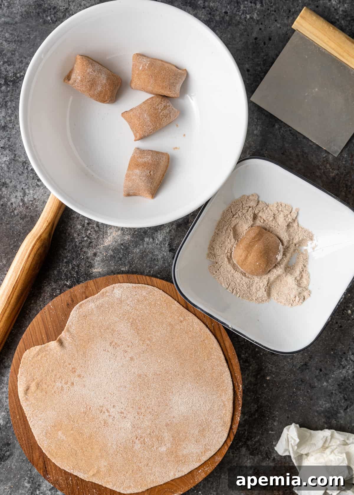 Overhead process shot displaying two white bowls with divided dough portions, next to a cutting board featuring a perfectly flat and thin circle of Roti dough, ready for cooking.