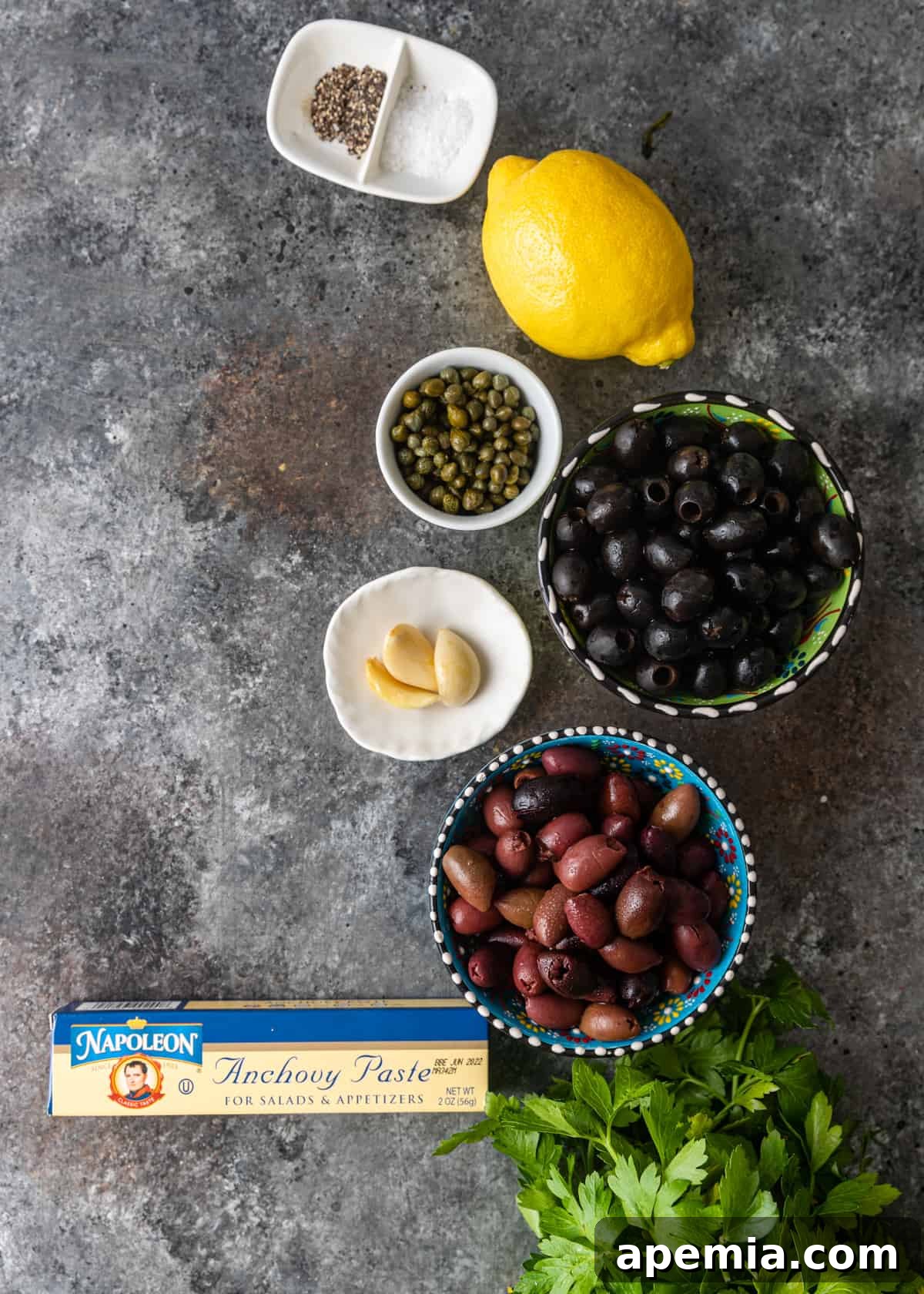 Savory Mediterranean Tapenade 4 Overhead view of various fresh ingredients laid out on a surface, including olives, parsley, garlic, capers, and a lemon, ready to make olive tapenade.