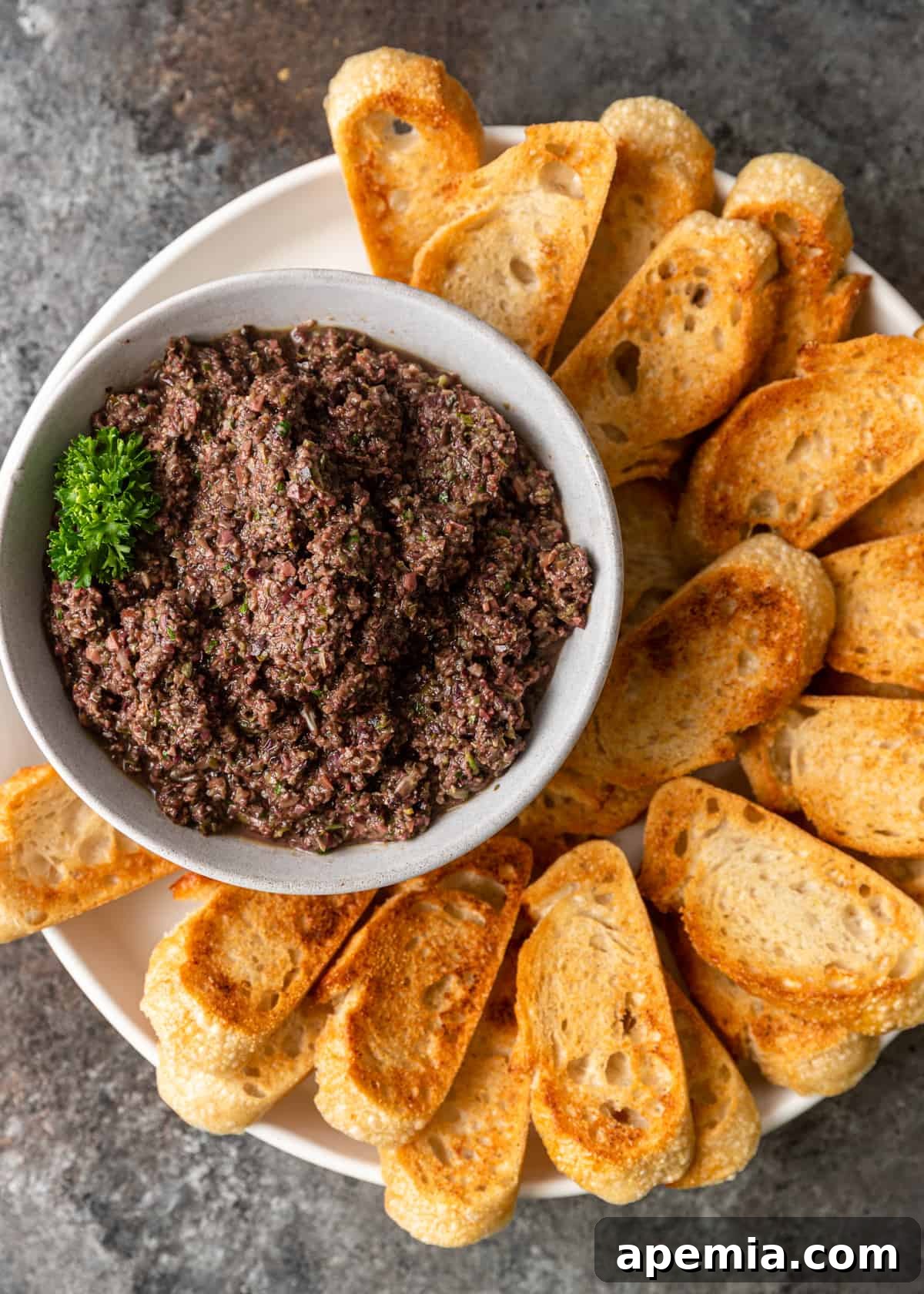 Savory Mediterranean Tapenade 8 Overhead view of a beautiful platter featuring a white bowl brimming with homemade olive tapenade, surrounded by neatly arranged golden-brown toasted baguette slices.
