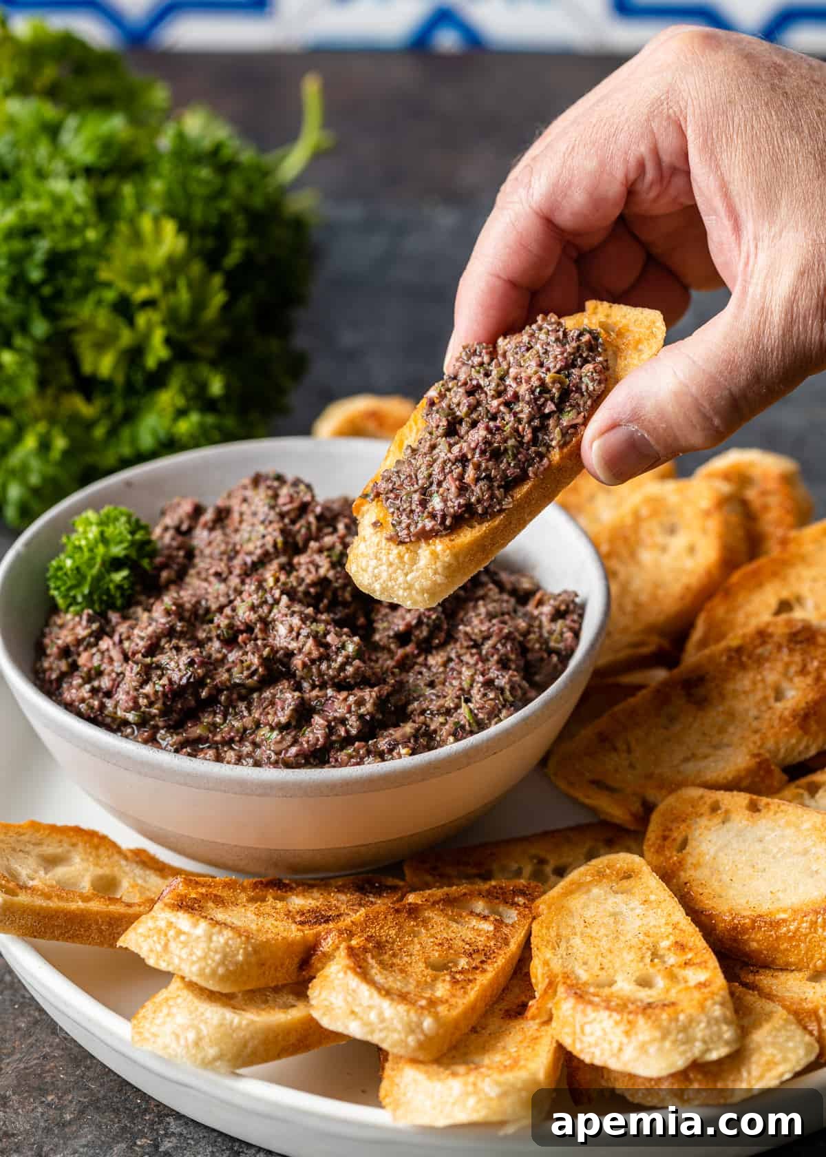 Savory Mediterranean Tapenade 10 Close-up side view of a hand holding a toasted baguette slice, dipping it into a bowl of homemade olive tapenade, with more baguette slices arranged on a white platter.