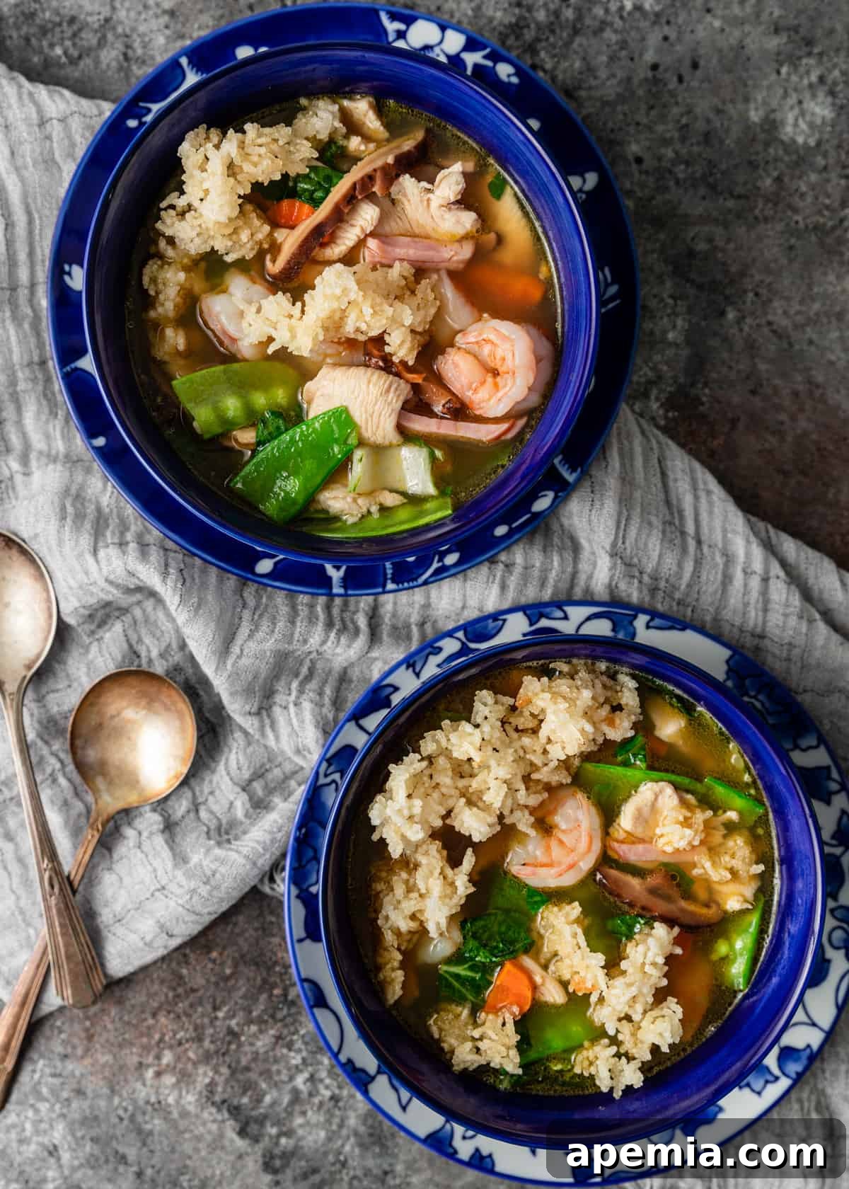 Overhead view of two bowls of Sizzling Rice Soup, elegantly arranged on a tea towel with two spoons, inviting you to share