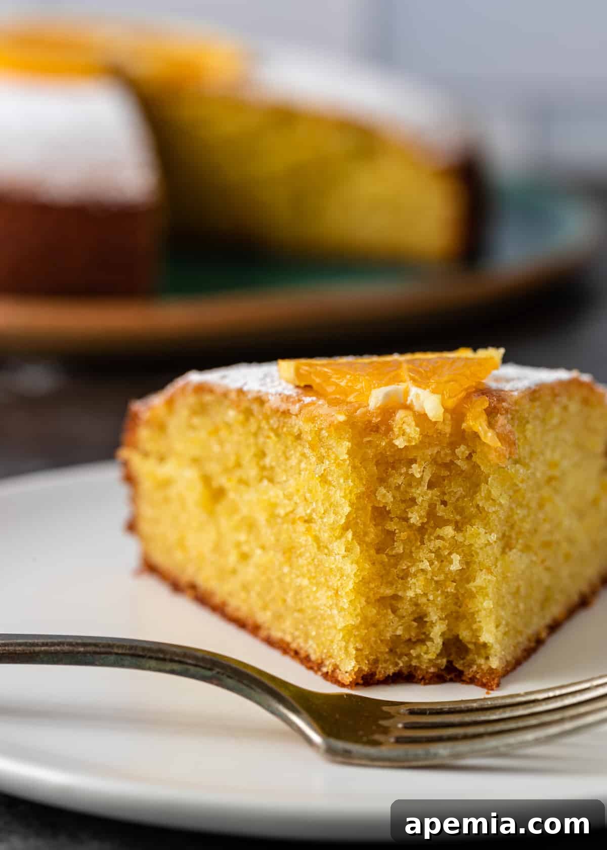 Closeup side view: a tempting slice of olive oil cake on a white plate, with a fork poised to take a bite, highlighting its texture and inviting appearance.