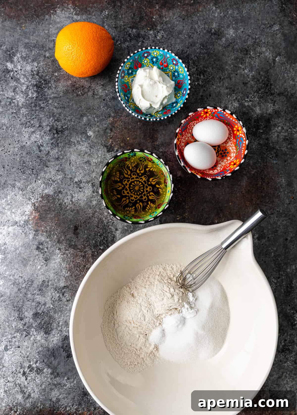 Overhead process shot: all the fresh ingredients perfectly arranged to make the olive oil cake recipe, including flour, sugar, eggs, olive oil, yogurt, and oranges.