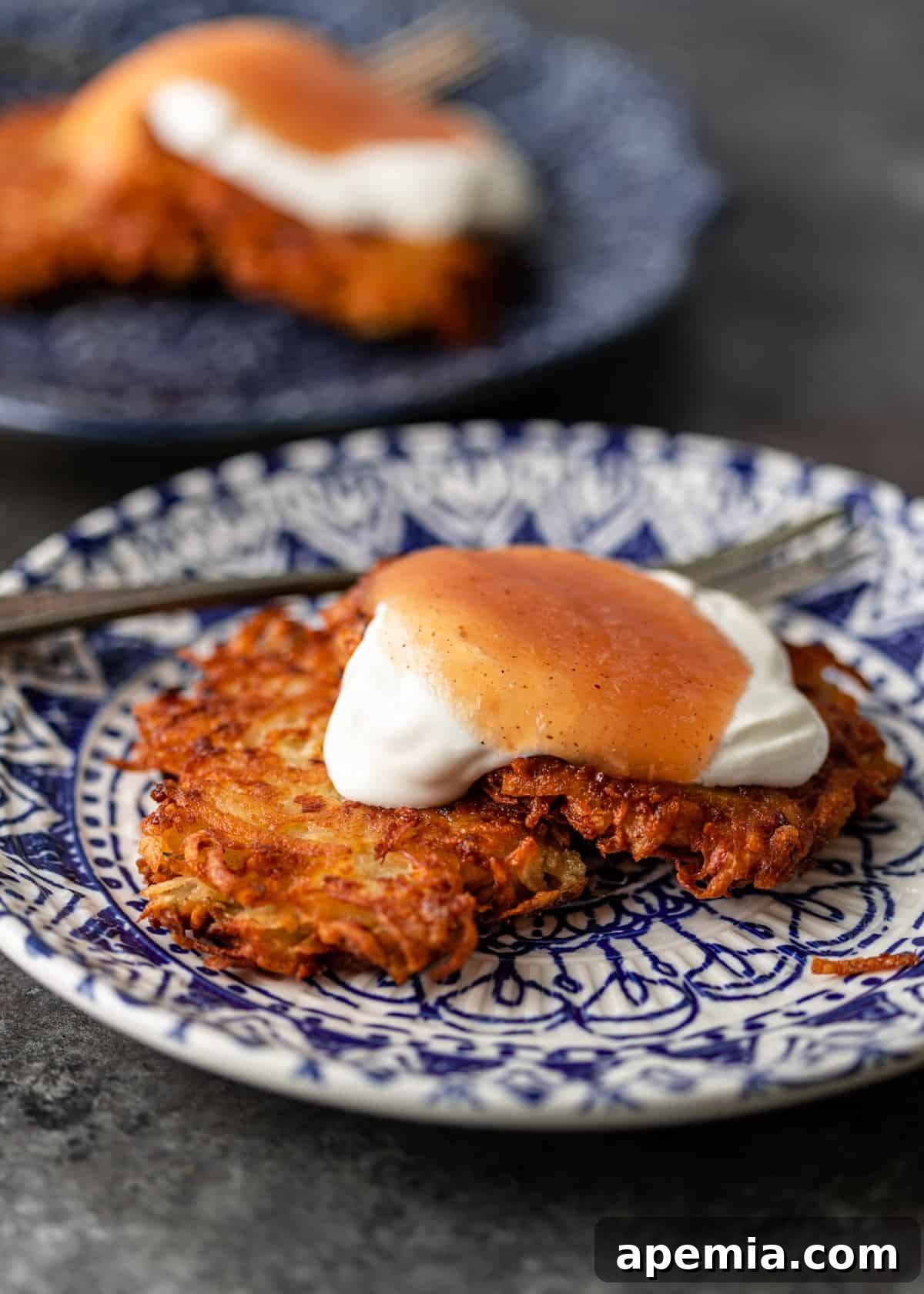 Closeup side view of two perfectly fried potato latkes on a white and blue plate, accompanied by a fork, ready to be enjoyed.