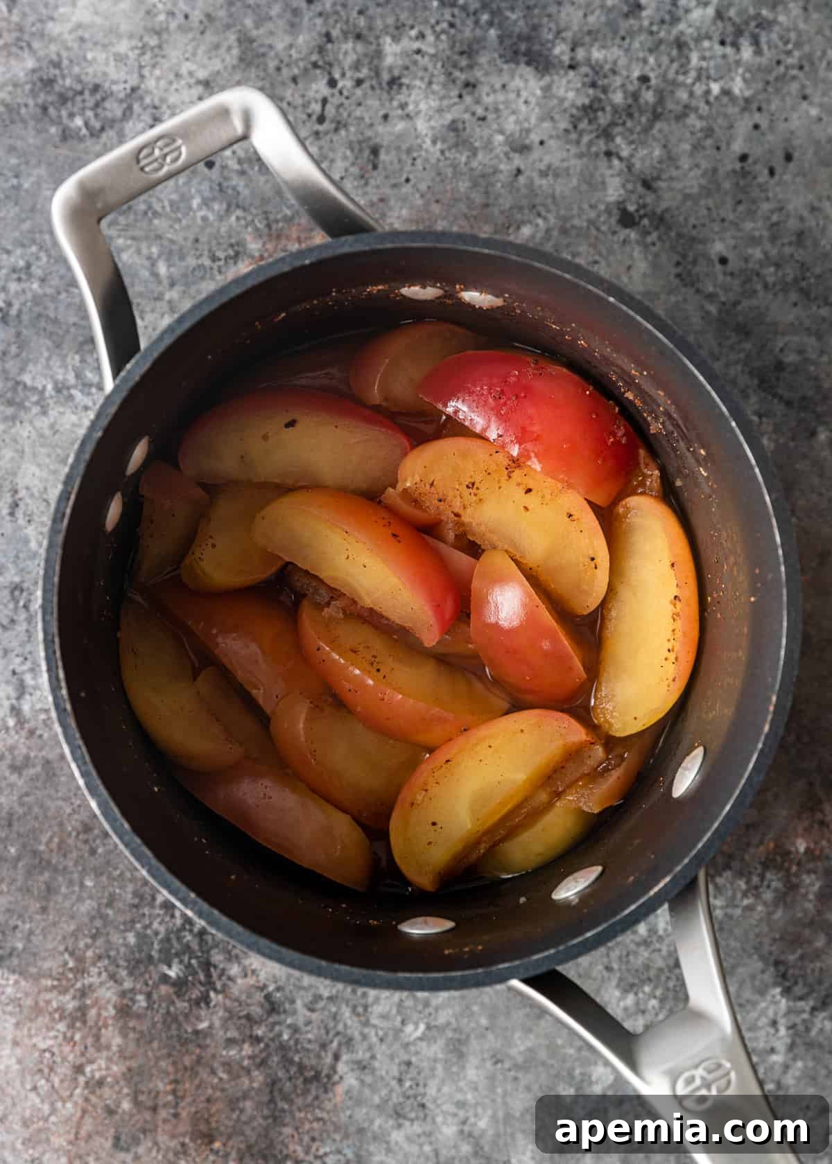 Overhead process shot showing apple slices simmering in a saucepan with sugar and spices, creating homemade applesauce.