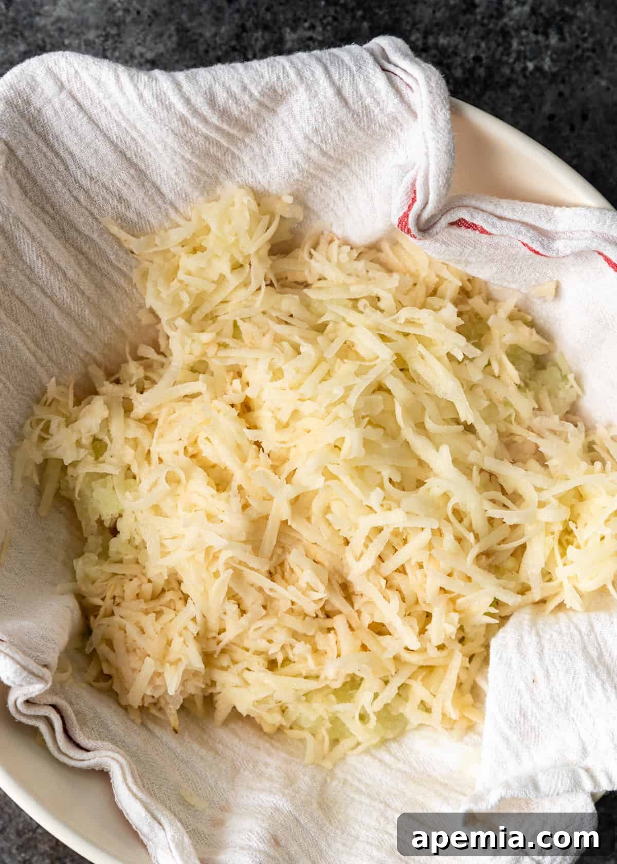 Overhead process shot illustrating the mixture of shredded potatoes and onion, placed onto a kitchen towel, ready for moisture removal.