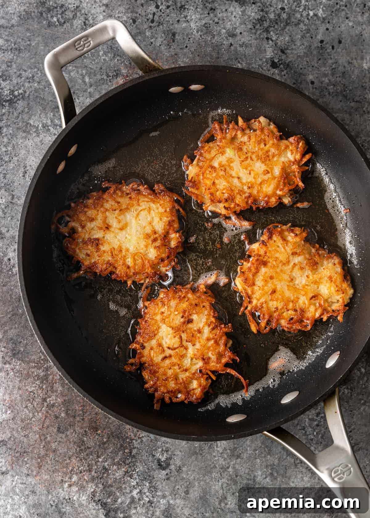 Overhead process shot of several potato latkes frying to a golden crisp in a hot skillet with shimmering oil.