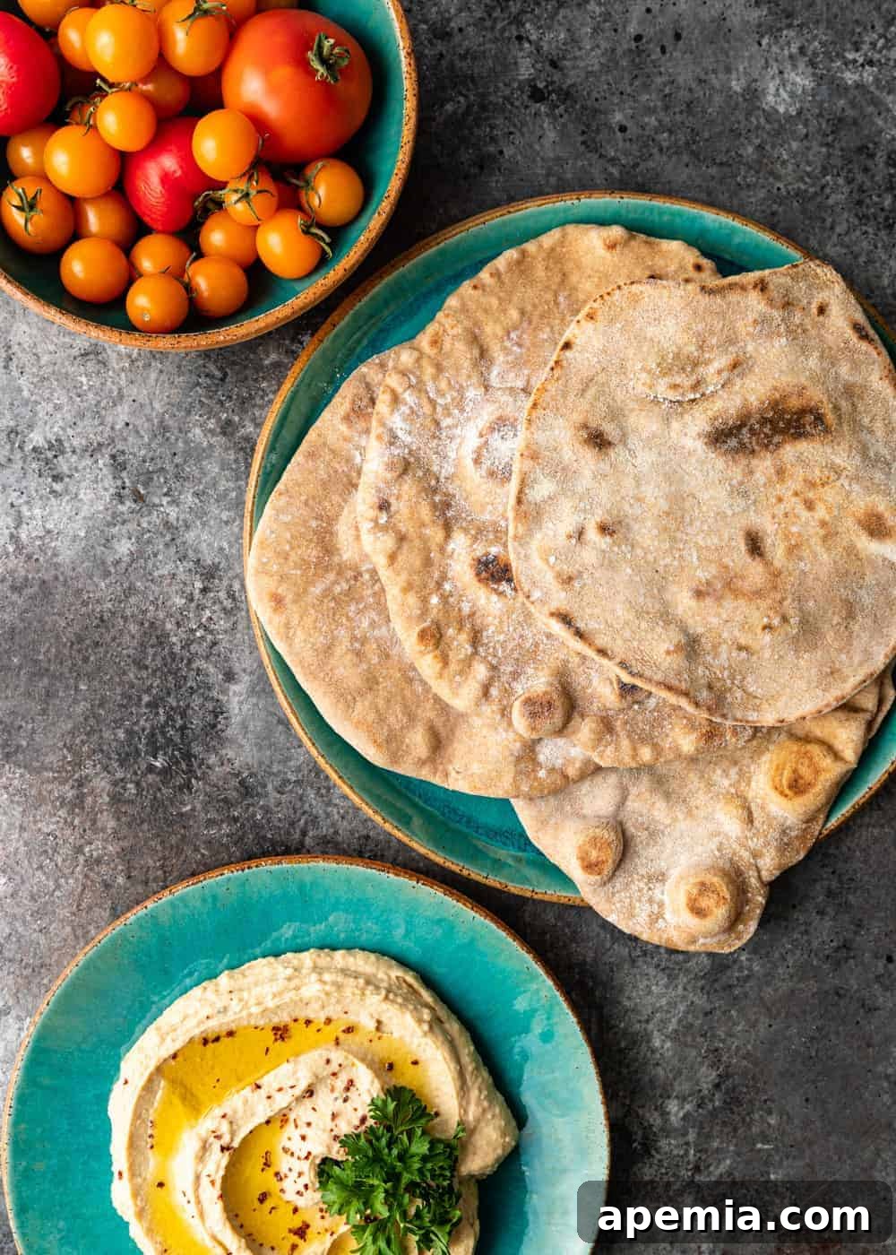 Overhead shot of a rustic ceramic plate showcasing freshly baked khubz (Arabic pita bread) artfully arranged next to a vibrant bowl of fresh tomatoes and a creamy plate of hummus drizzled with olive oil, inviting a delightful culinary experience.