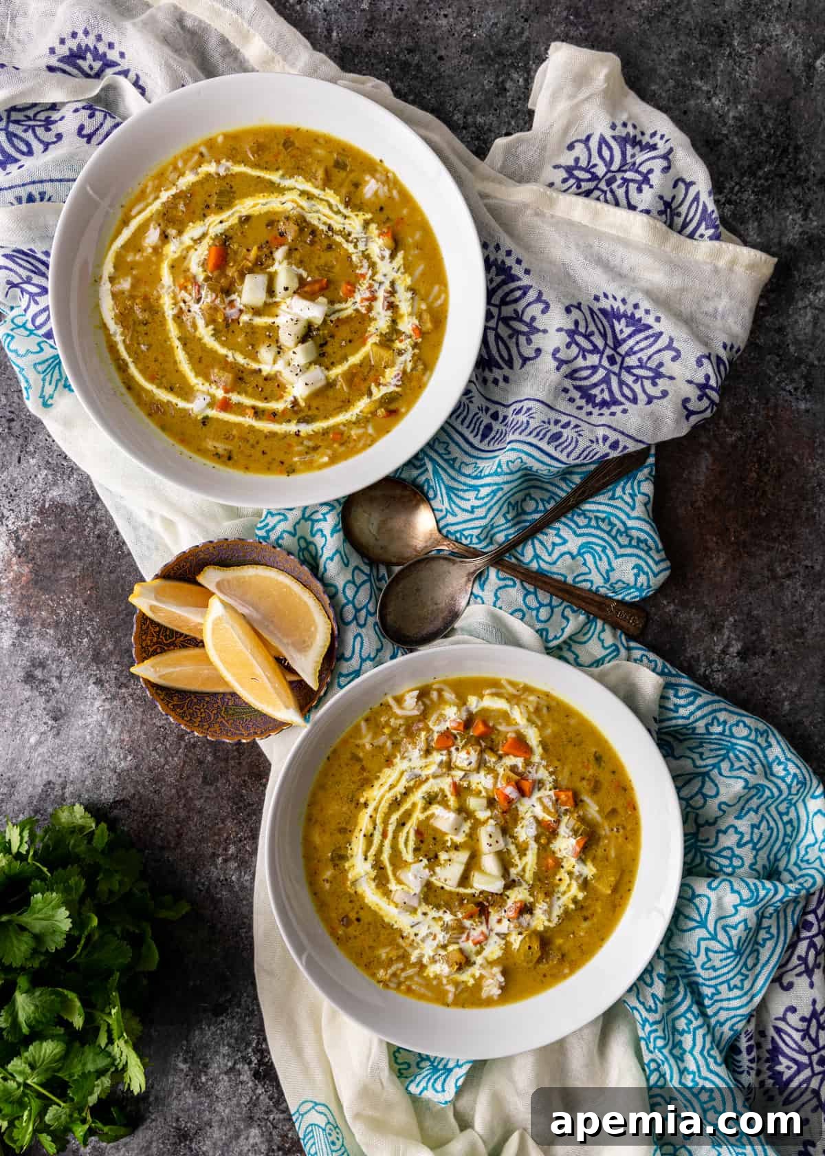 overhead: two white bowls of mulligatawny soup on tea towels with two spoons and sliced lemons on dish