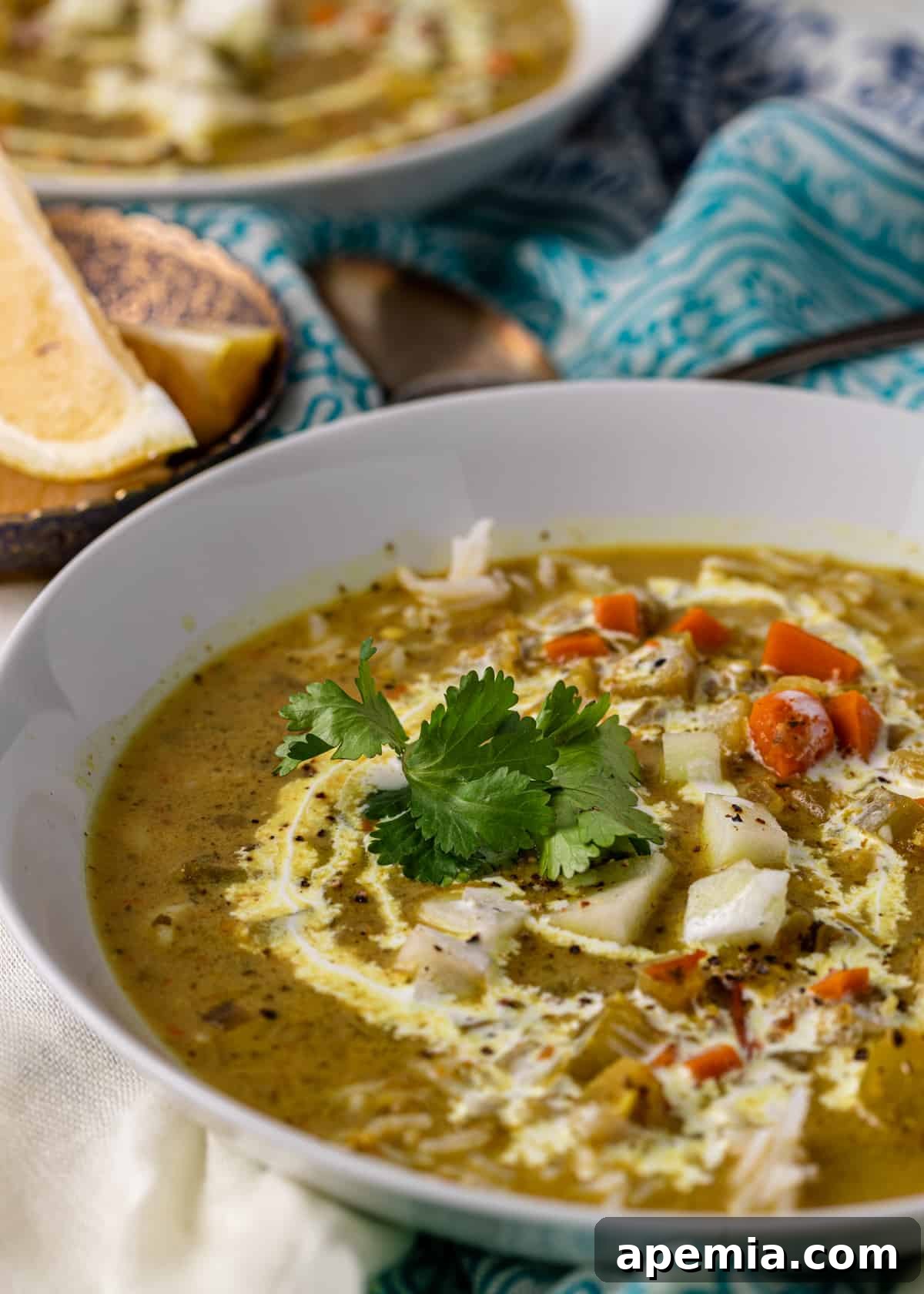 closeup side view: mulligatawny soup in a white bowl with tea cloth off to side with two spoons and lemon slices on dish