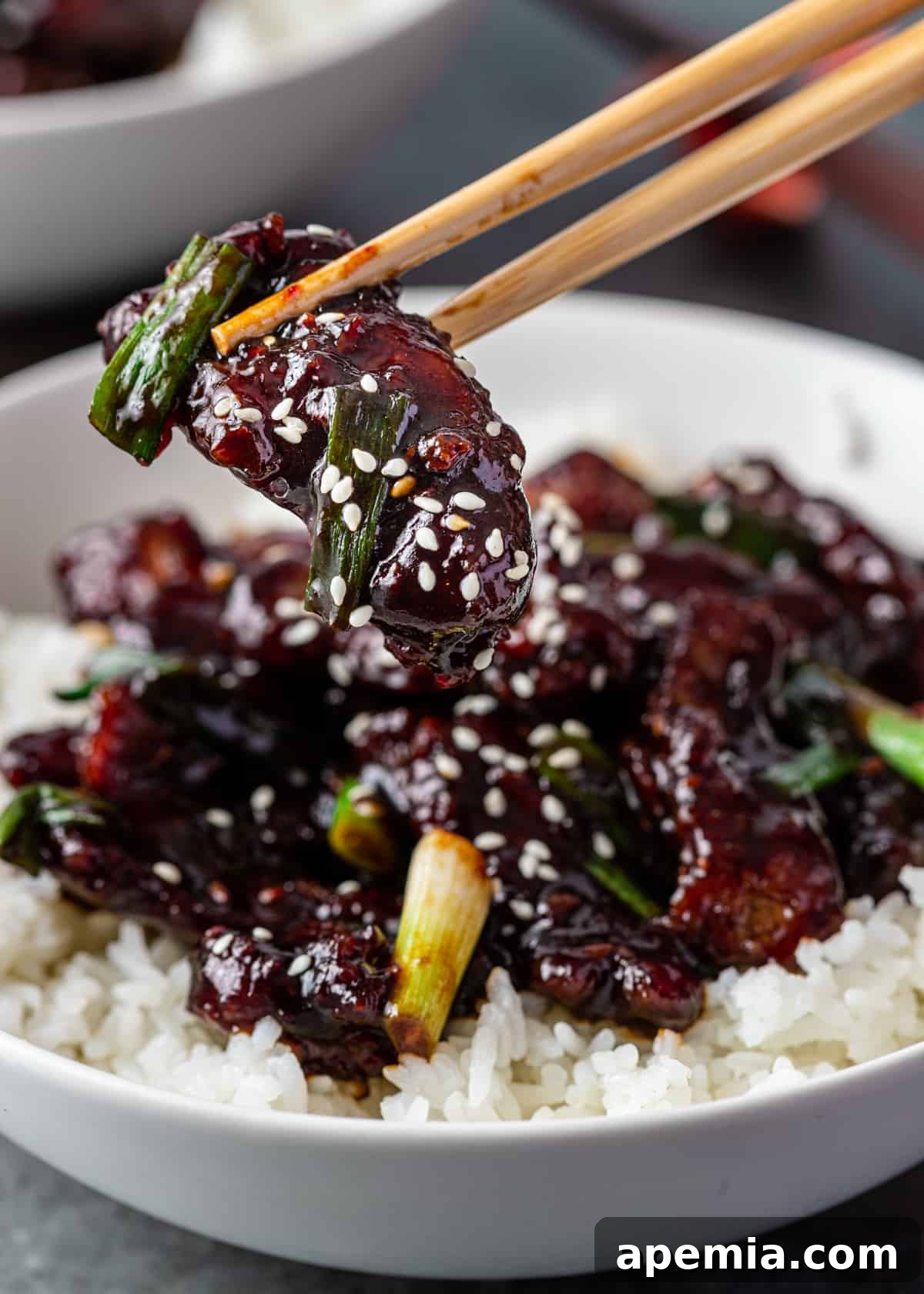 closeup side view: mongolian beef in chopsticks overtop of white bowl with rice & mongolian beef stir fry