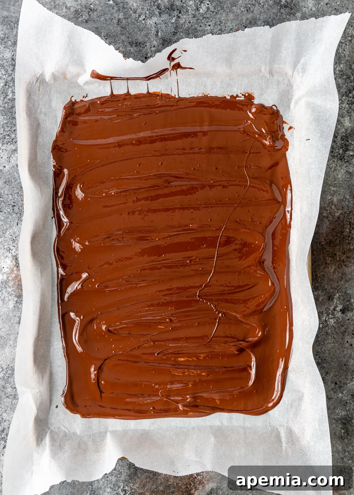 Overhead process shot demonstrating how to spread melted dark chocolate evenly onto a parchment-lined baking sheet.