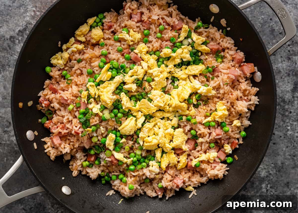 Overhead shot of green peas, chopped green onions, and scrambled eggs being added to the pork fried rice in a wok.