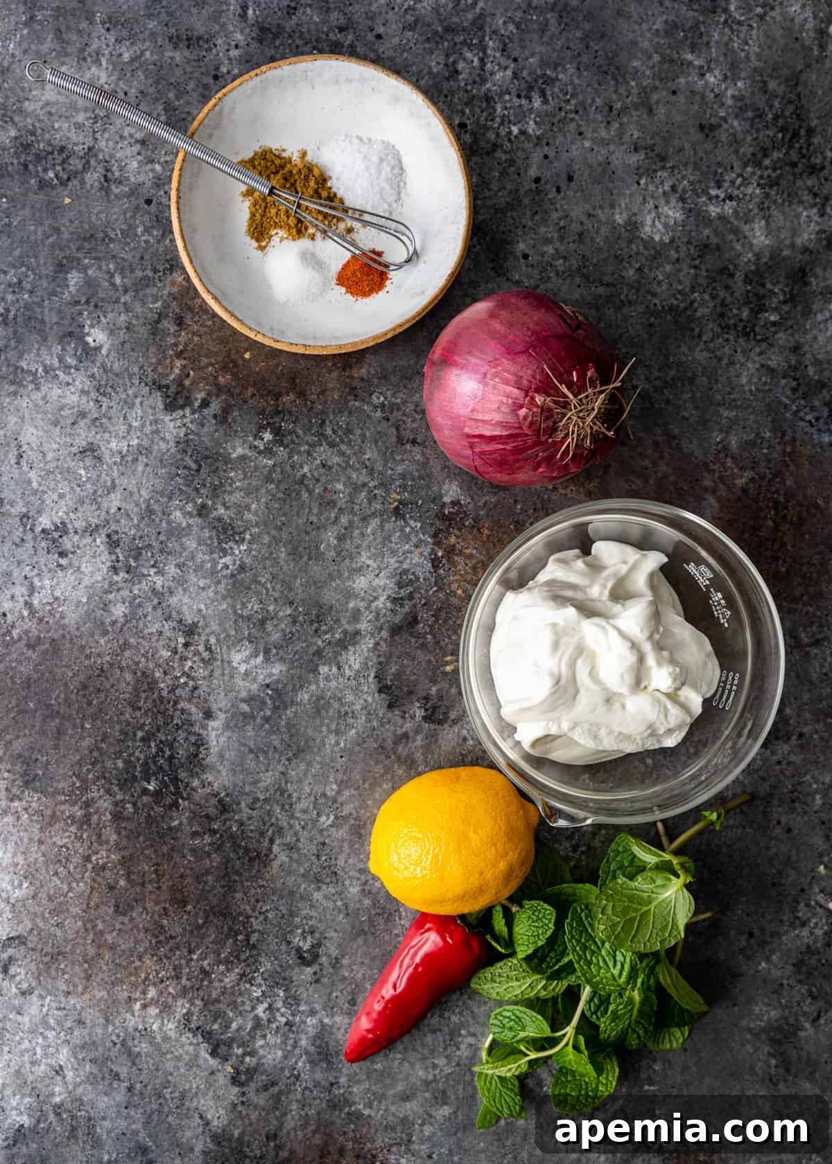 Various fresh ingredients laid out on a wooden surface, including Greek yogurt, lemons, red onions, chilis, and mint leaves, prepared for making Onion Raita.